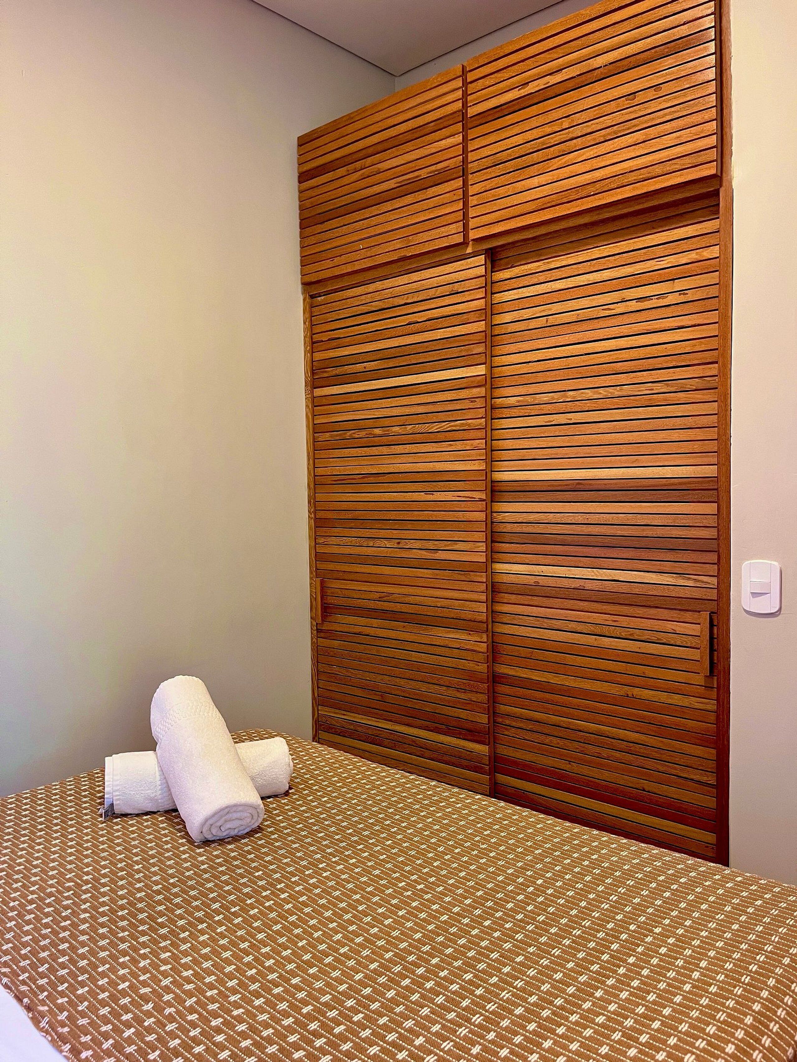A bedroom with a bed, a wooden wardrobe, and two rolled towels on the bed.