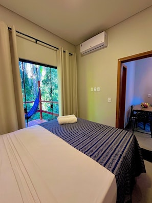 A bedroom with a bed, a window with curtains, an air conditioning unit, and a view of greenery outside.