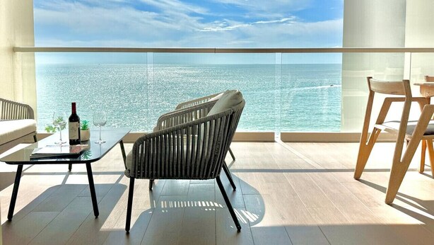 Un balcón con vistas al mar, una mesa con una botella y vasos, y dos sillas.