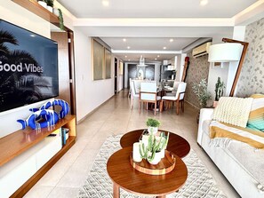 Ein modernes Wohnzimmer mit Flachbildfernseher, einem Couchtisch mit Sukkulenten als Mittelstück, einer Couch mit Überwurfdecke und einem Essbereich im Hintergrund.
