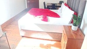 A bedroom with a bed, a wooden floor, a ceiling fan, a TV, a door, and a small table with chairs.