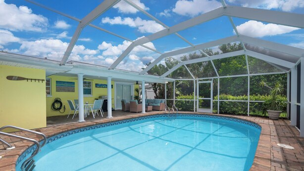 An indoor swimming pool with a glass ceiling, surrounded by a patio area with seating and a view of greenery. - Sunshine Paradise Minutes From Action. Grill, Bikes, Beach Gear & More Included! (Cape Coral)