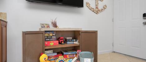 A room with a TV mounted on the wall, a wooden cabinet with toys, a toy chicken, and a trash bin labeled "Kids Toys".