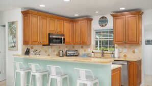 A kitchen with wooden cabinets, a microwave, a toaster, and a dishwasher. There are three white bar stools and a window with a view of greenery. - Sunshine Paradise Minutes From Action. Grill, Bikes, Beach Gear & More Included! (Cape Coral)