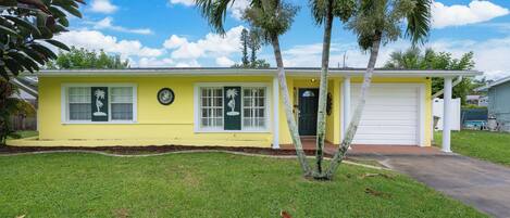 A yellow single-story house with a front porch, palm trees, and a garage.