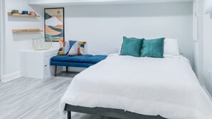 Een slaapkamer met een bed, een blauwe bank, een witte nachtkastje en een wandplank.