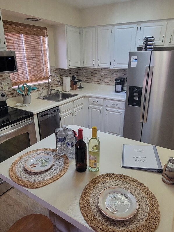 A modern kitchen with white cabinets, a stainless steel refrigerator, and a dining area with woven placemats and a wine bottle.