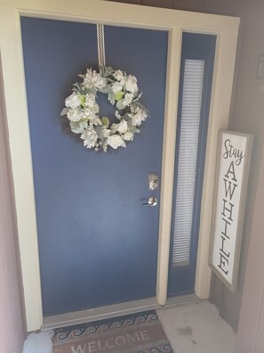 A blue door with a white wreath, a doorbell, and a 'Stay Awhile' sign.