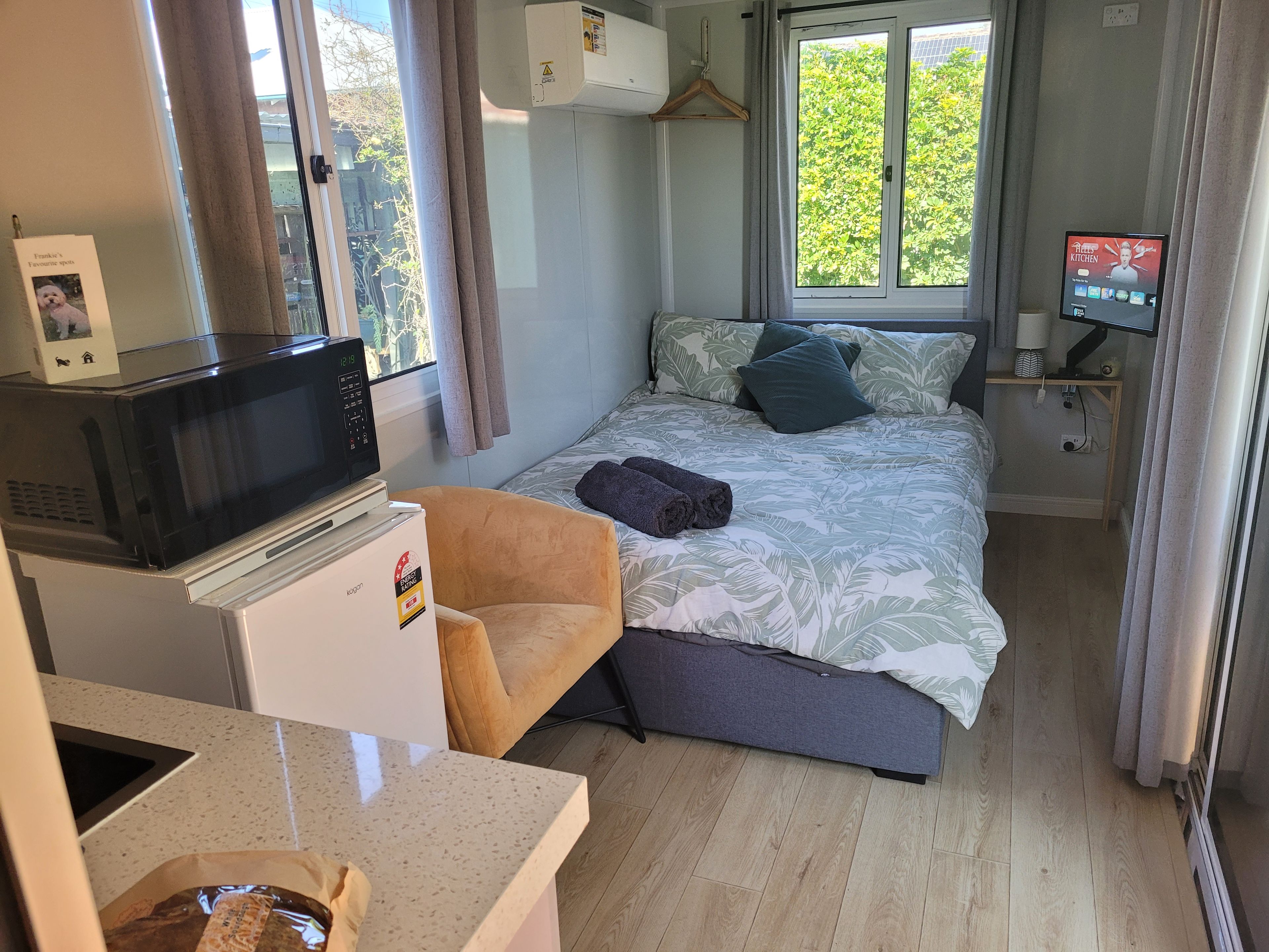 A compact hotel room with a bed, a small desk with a microwave, a chair, and a TV on a stand.
