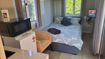 A compact hotel room with a bed, a small desk with a microwave, a chair, and a TV on a stand.