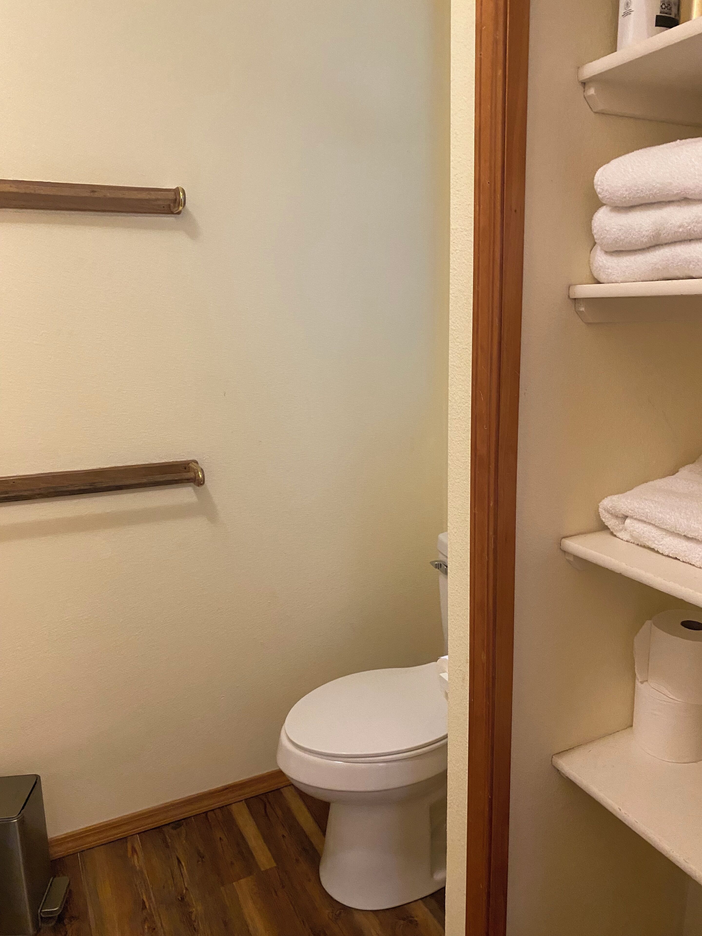 Un baño con inodoro, suelo de madera y paredes blancas. Hay dos toalleros y un estante con toallas.