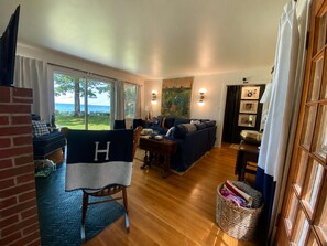 A cozy living room with a large window, a sofa, a chair with a blanket, a wooden floor, and a basket.