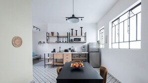 A modern kitchen with a dining area, featuring a black table with chairs, a bowl of fruit, and a hanging light fixture.