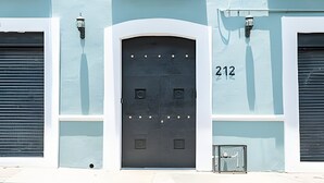 A building with a large arched door marked with the number 212, flanked by two closed shutters and a small window with a metal grate.