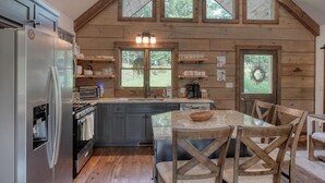 A rustic cabin-style kitchen with a large refrigerator, a stove, and a sink. There are wooden chairs around a table, and the kitchen has a high ceiling with large windows. - Kenny's Place, Suches (Suches)