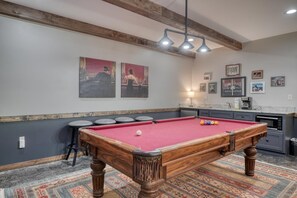Une table de billard recouverte d’un feutre rouge, un coin bar avec des tabourets et un espace cuisine équipé d’un four à micro-ondes.