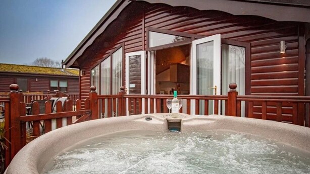 A hot tub in front of a wooden cabin with a wooden deck and a glass door leading to the interior. - A lodge that sleeps 4 guests  in 2 bedrooms (Windermere)