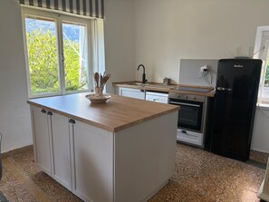 Cozinha com ilha central, geladeira preta e janela com vista para o verde.