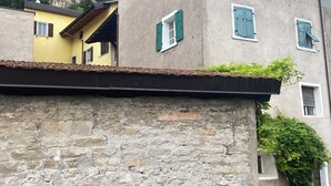 A stone wall with a tiled roof, a building with a yellow facade and green shutters, and a mountain in the background.