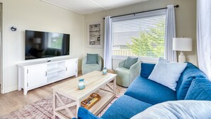 Una sala de estar con televisor, un mueble blanco, sofá azul y una ventana con persianas.