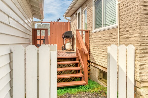A wooden deck leading to a house with a white picket fence and a covered area with a grill.