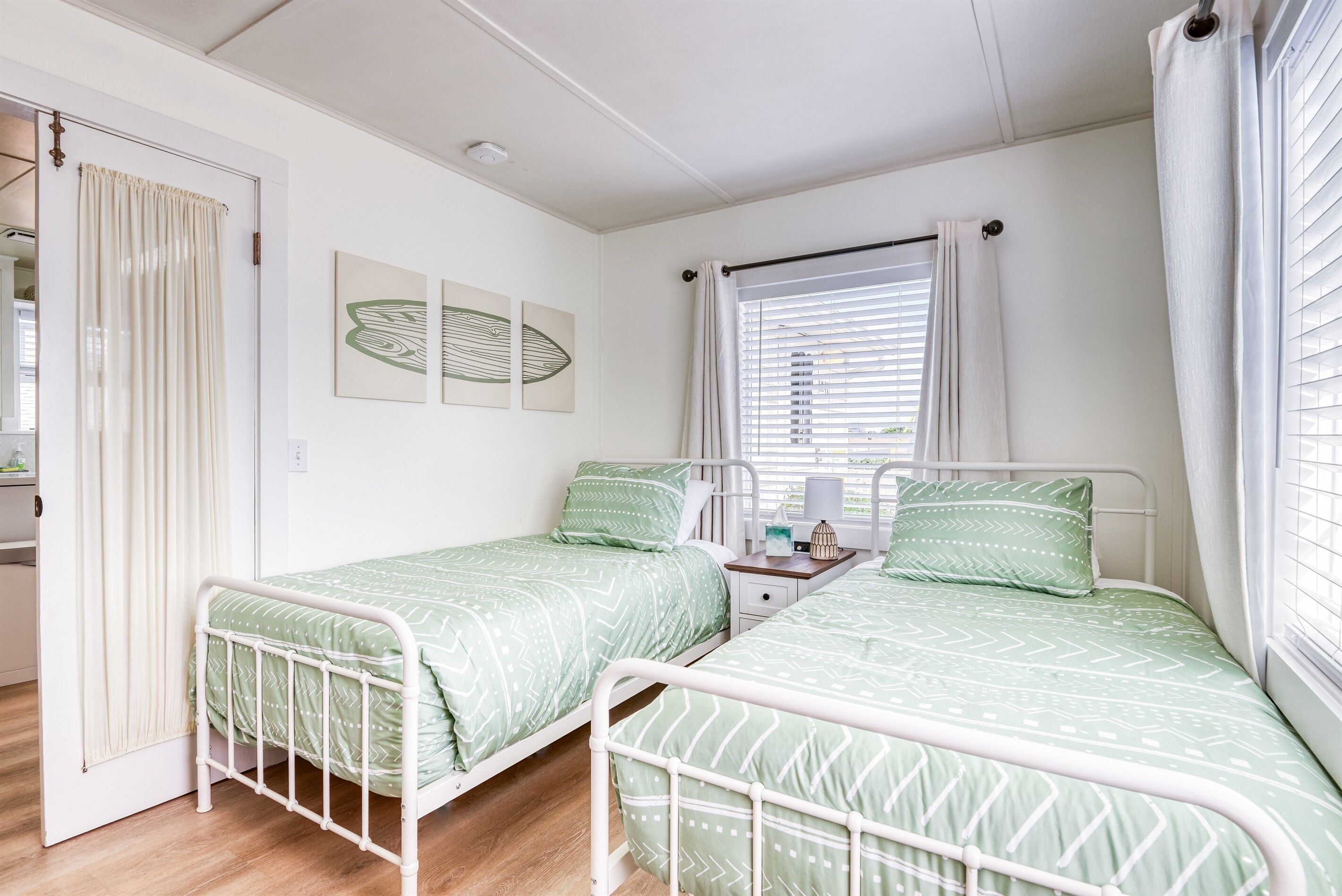 A twin bedroom with two beds, a window with blinds, a bedside table, and a door.