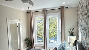 A bedroom with a bed, a ceiling fan, two windows with curtains, a door, and a lamp. - Remodeled Old-World Home, Walkable to Rino, Ballpark,& Downtown (Denver)