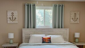 Une chambre à coucher comprenant un lit, un ventilateur de plafond, deux tables de chevet, une fenêtre avec des rideaux et deux éléments de décoration fixés au mur.