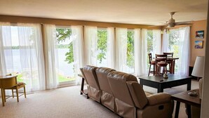 Un salon comprenant un canapé, une table à manger avec des chaises et un ventilateur de plafond. De grandes fenêtres, ornées de rideaux légers, offrent une vue sur l’extérieur.