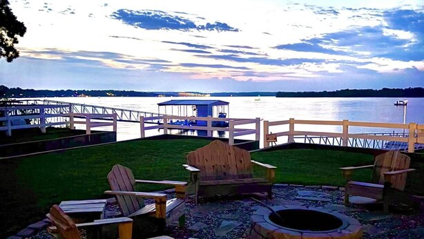Une terrasse au bord de l’eau, dotée de fauteuils Adirondack et d’un foyer, offrant une vue sur un lac paisible et un pont situé au loin.
