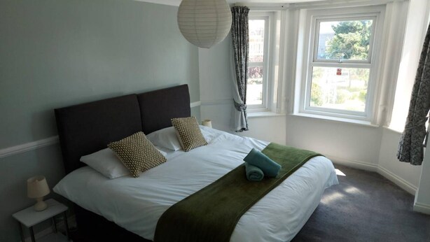 A neatly made bed with white linens and two patterned pillows, a dark headboard, and a green throw blanket. There is a bedside lamp on a small white nightstand. - in Bournemouth With 2 Bedrooms and 1 Bathrooms (Bournemouth)
