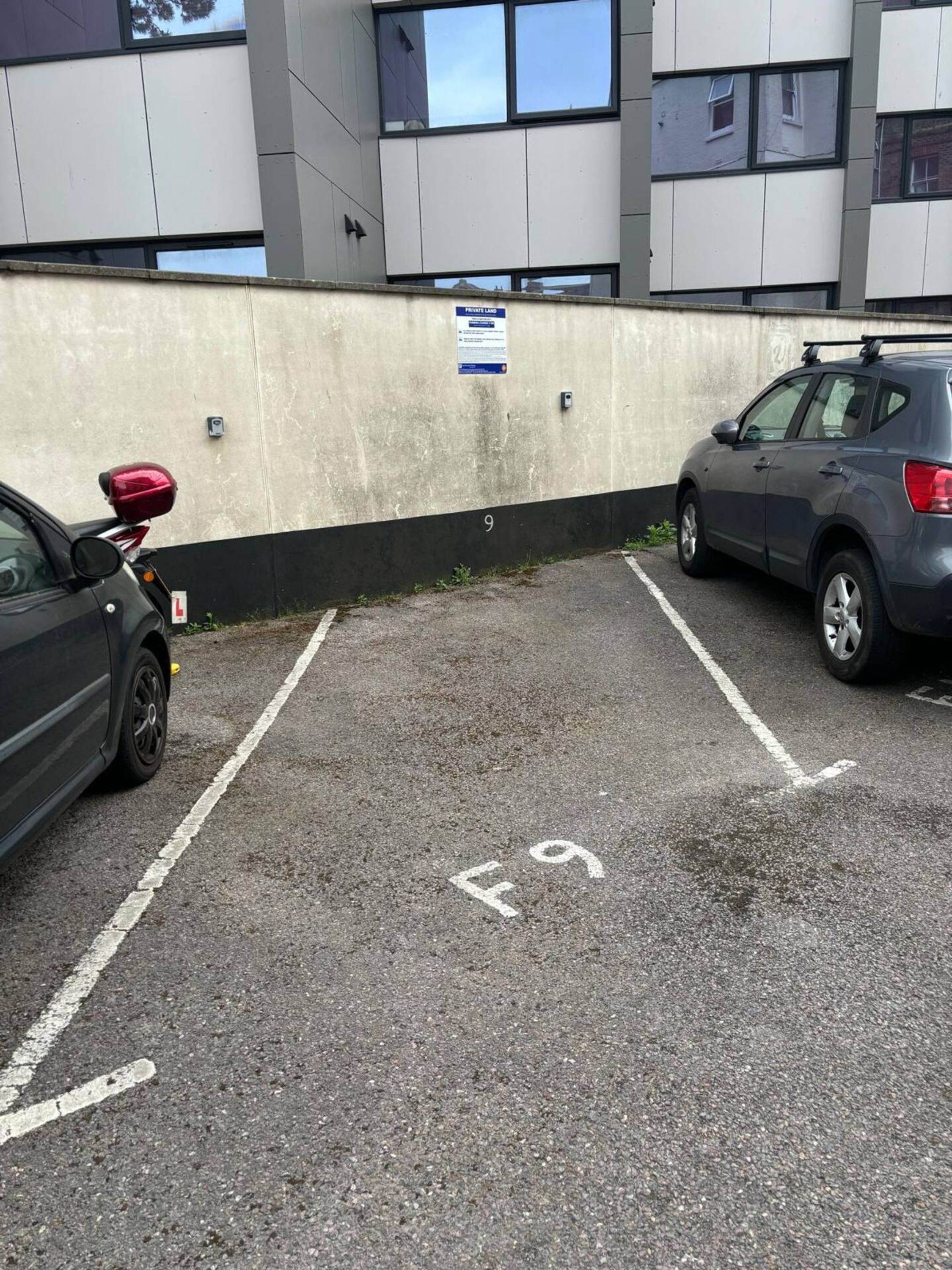 Un estacionamiento con espacios delimitados, un muro de concreto y un edificio con ventanas al fondo.