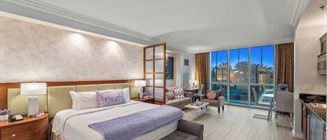 A spacious hotel room with a large bed, a desk, a chair, and a view of the city through large windows.