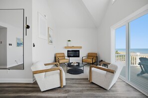 Ein Zimmer mit Fernseher, zwei Sesseln, einem kleinen Couchtisch und Blick auf den Strand.