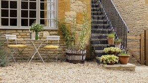 A stone building with a white window, a wooden bench, a small table, and a metal chair. There are potted plants and a gravel ground. - Beautifully presented dog friendly - The Coach House at Broadwell Manor (Broadwell)
