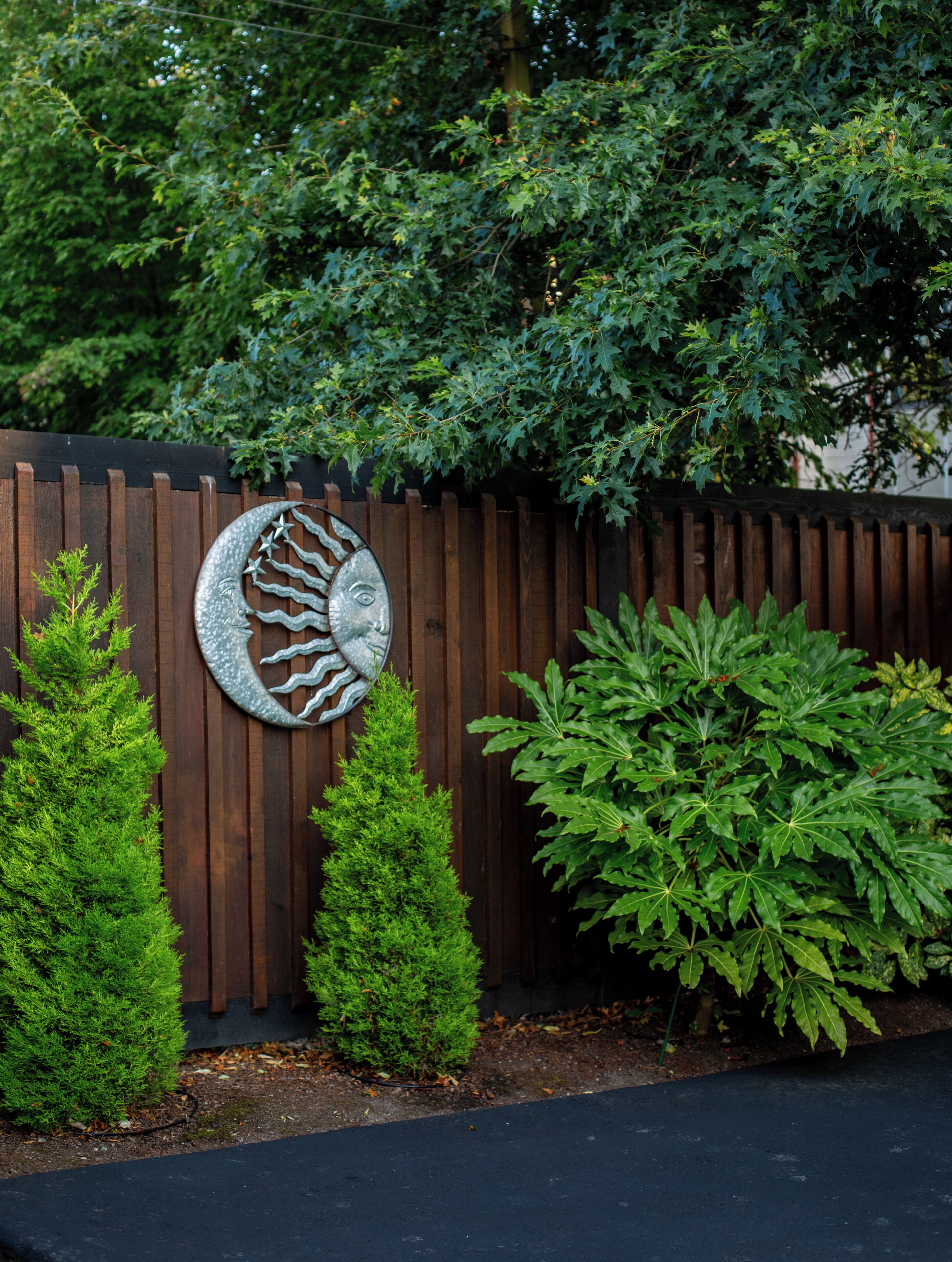 A wooden fence with a decorative metal piece, two small coniferous trees, and a larger leafy shrub.
