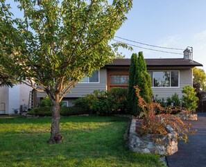 A two-story house with a stone retaining wall, a tree in the front yard, and a clear sky.