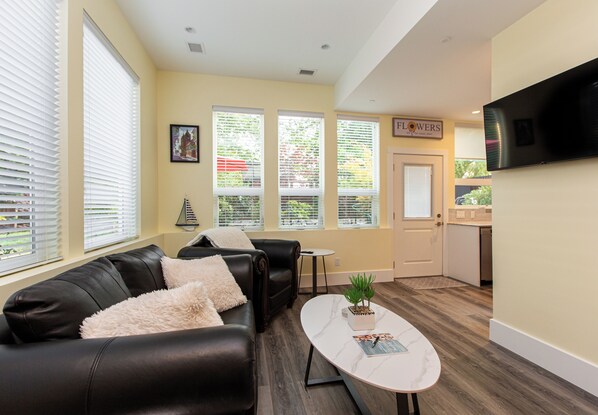 A modern living room with a black leather sofa, a white coffee table, and a potted plant. There are large windows with blinds, a flat-screen TV mounted on the wall, and a door labeled "Flowers".