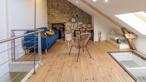 Een loft-achtige kamer met een stenen muur, houten balken, een blauwe bank, een rieten stoel en een dakraam.