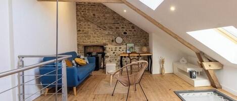 Une pièce de style loft avec un mur en pierre, des poutres en bois, un canapé bleu, un fauteuil en osier et une lucarne.