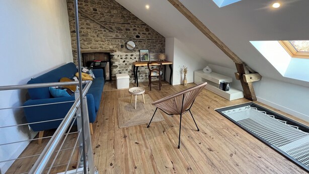 Een loft-achtige kamer met een schuine kap, een stenen muur, houten balken, een blauwe bank, een rieten stoel en een dakraam.