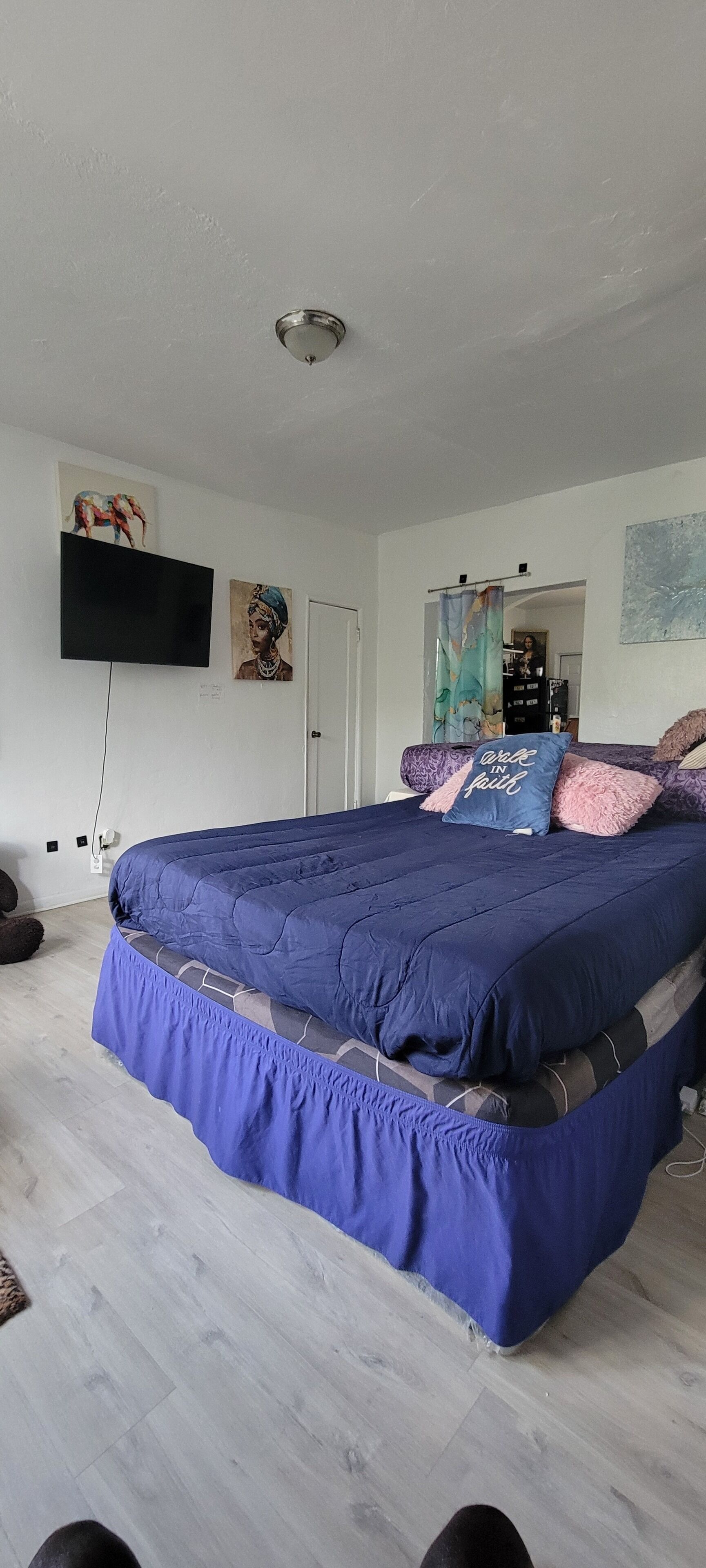 A bedroom with a large bed, a flat-screen TV mounted on the wall, and a door leading to another room.
