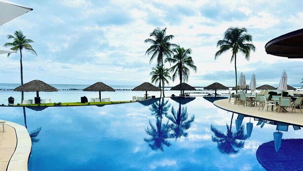 Una piscina infinita azul cristalina con sillas de descanso y sombrillas, palmeras y vistas al océano.