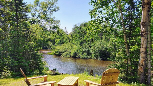 Dos sillas de madera tipo Adirondack y una pequeña mesa instaladas en un césped con vista a un río rodeado de denso bosque.