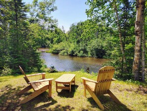 Dos sillas de madera tipo Adirondack y una pequeña mesa instaladas en un césped con vista a un río rodeado de denso bosque.