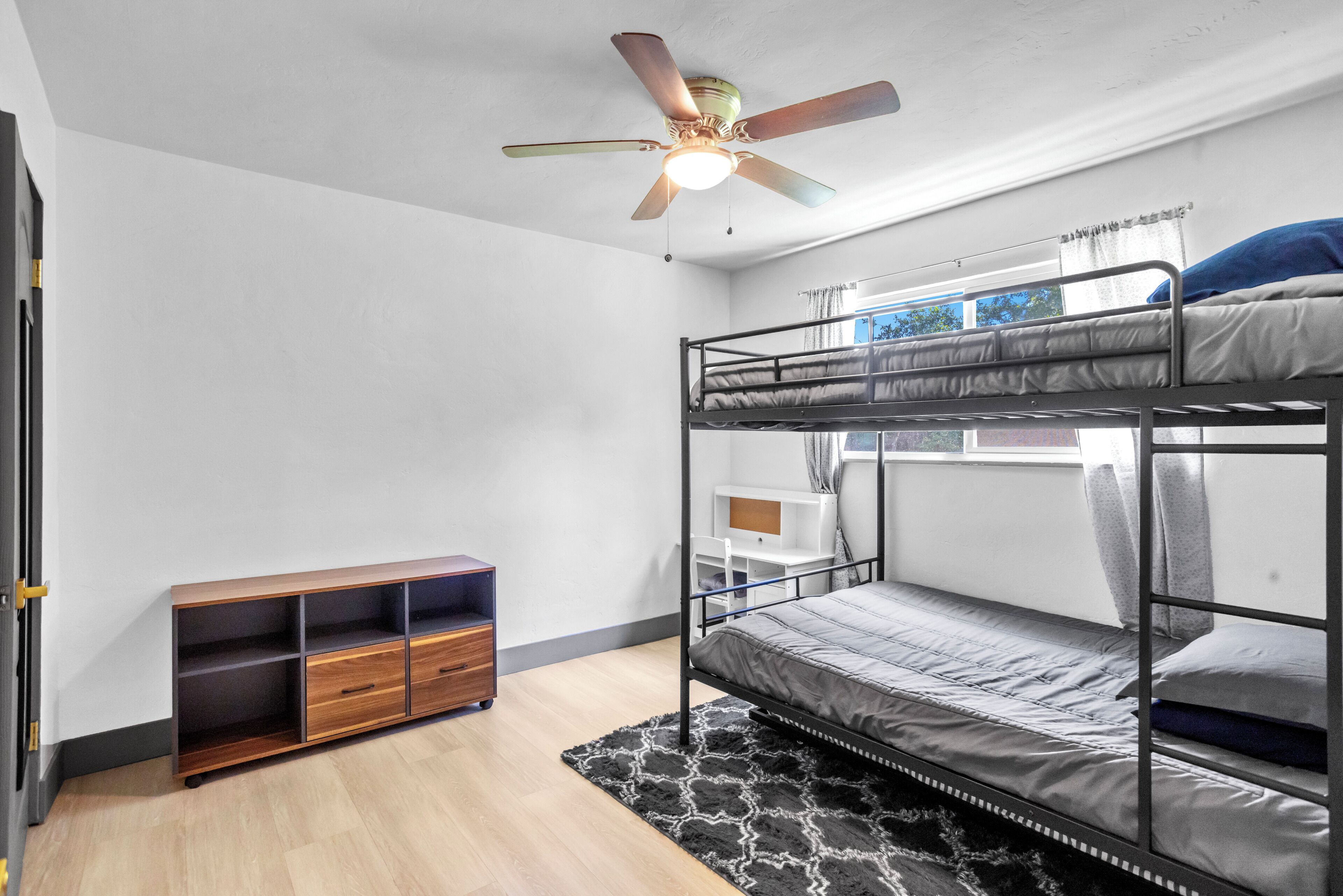 A bunk bed room with a ceiling fan, a wooden cabinet, and a window with curtains.