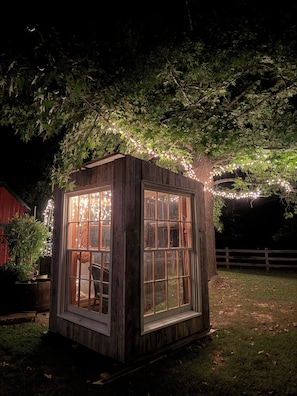 Une petite construction en bois dotée de grandes fenêtres, entourée de verdure et illuminée de guirlandes lumineuses.