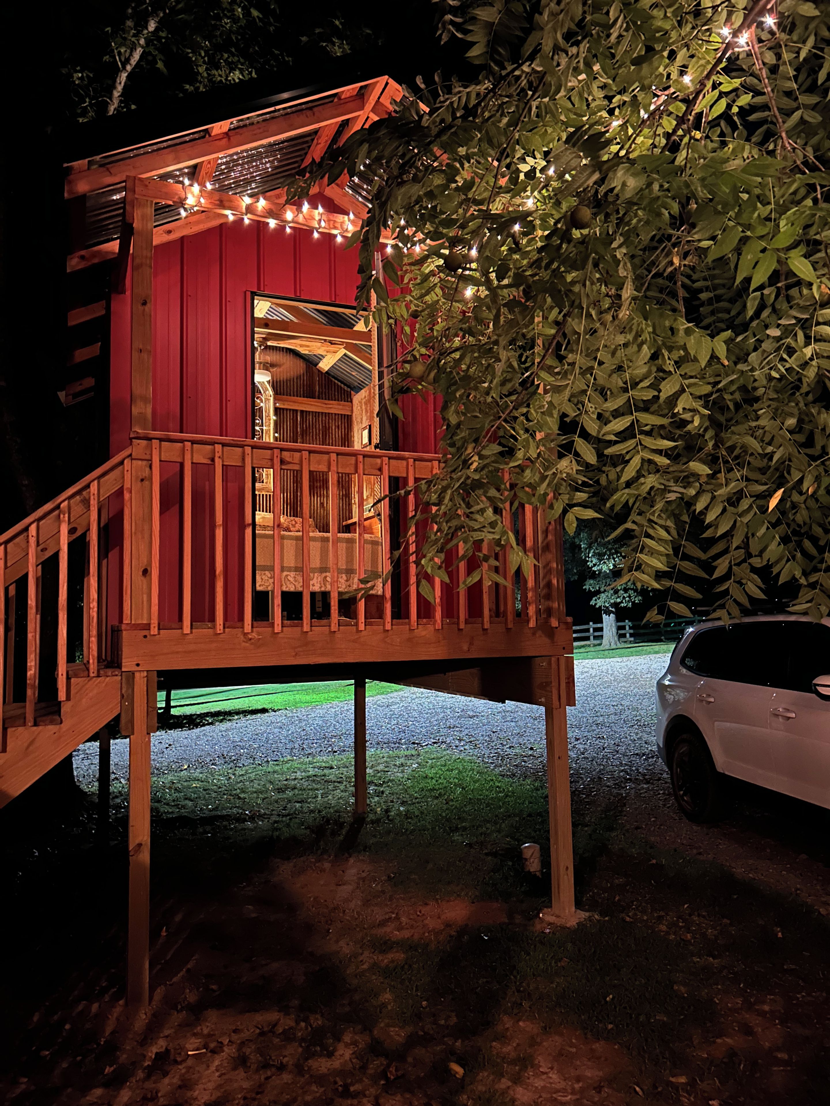 A wooden deck structure with a red exterior and string lights, a car parked nearby, and a tree in the foreground.