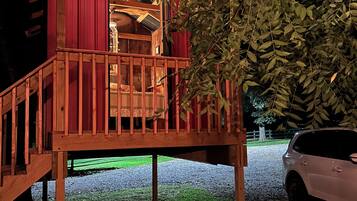A wooden deck structure with a red exterior and string lights, a car parked nearby, and a tree in the foreground.