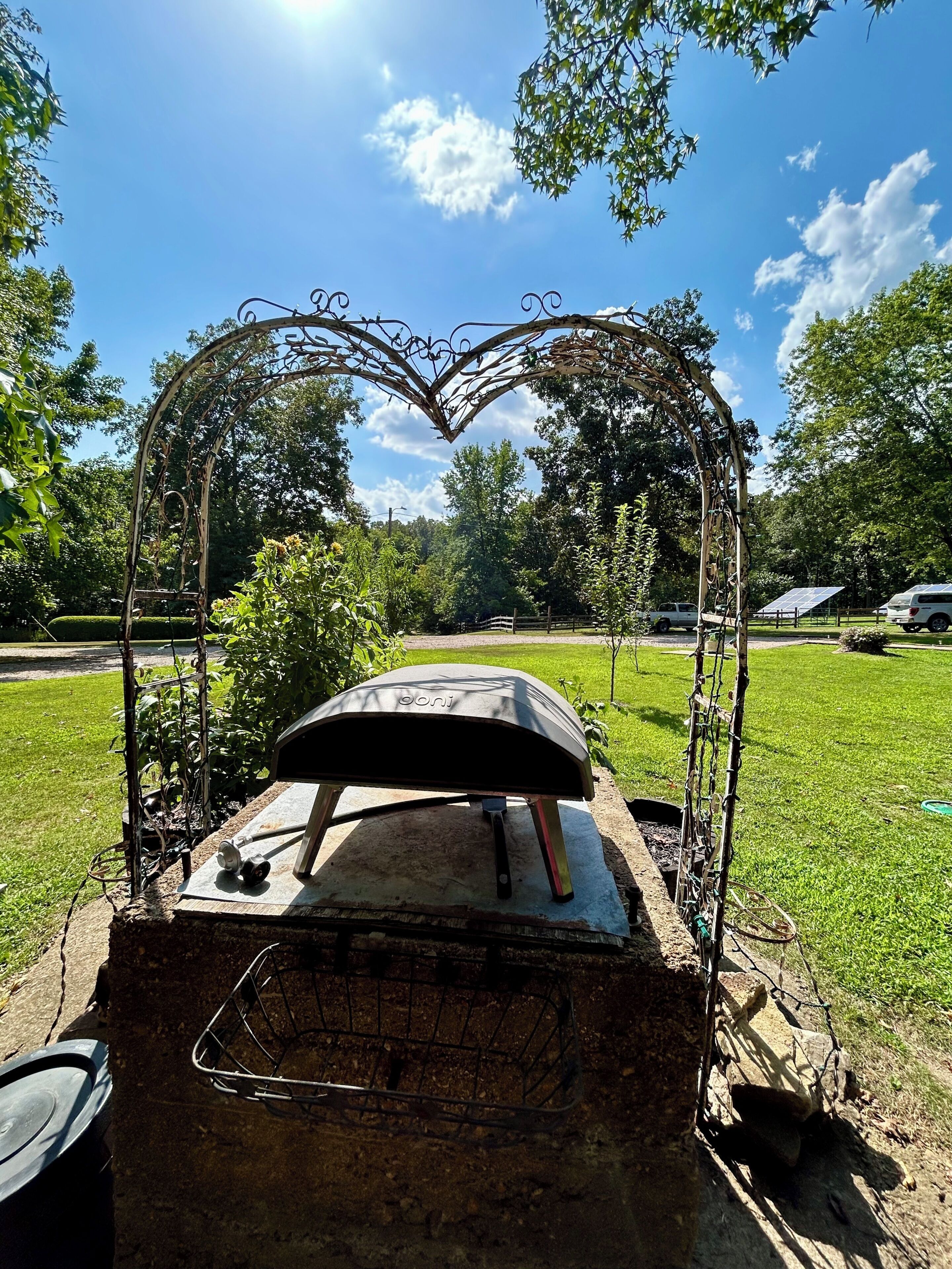 An outdoor pizza oven with a heart-shaped metal arch, surrounded by greenery and parked vehicles.